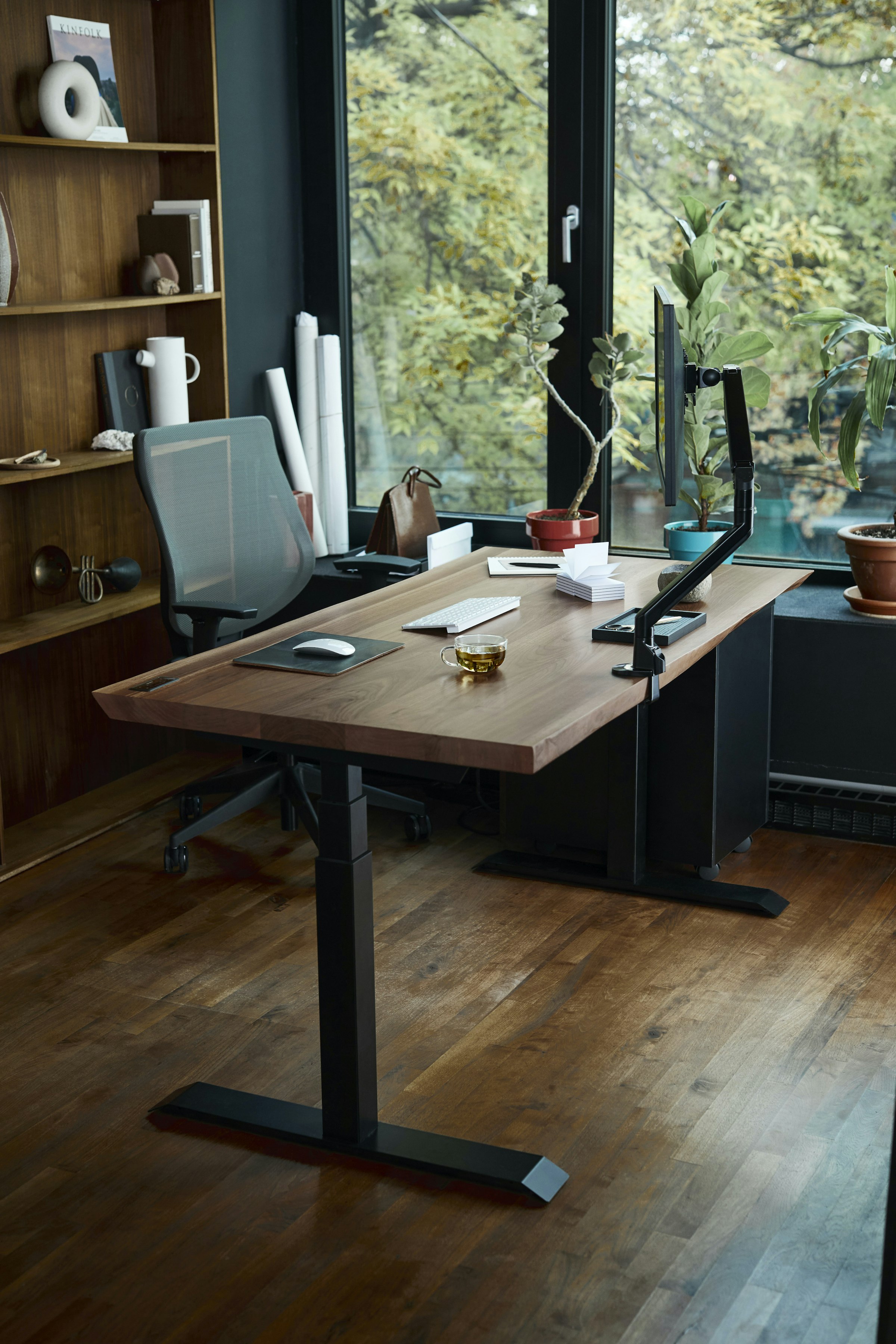 A laptop on a wooden desk in front of a window, with plants and natural daylight