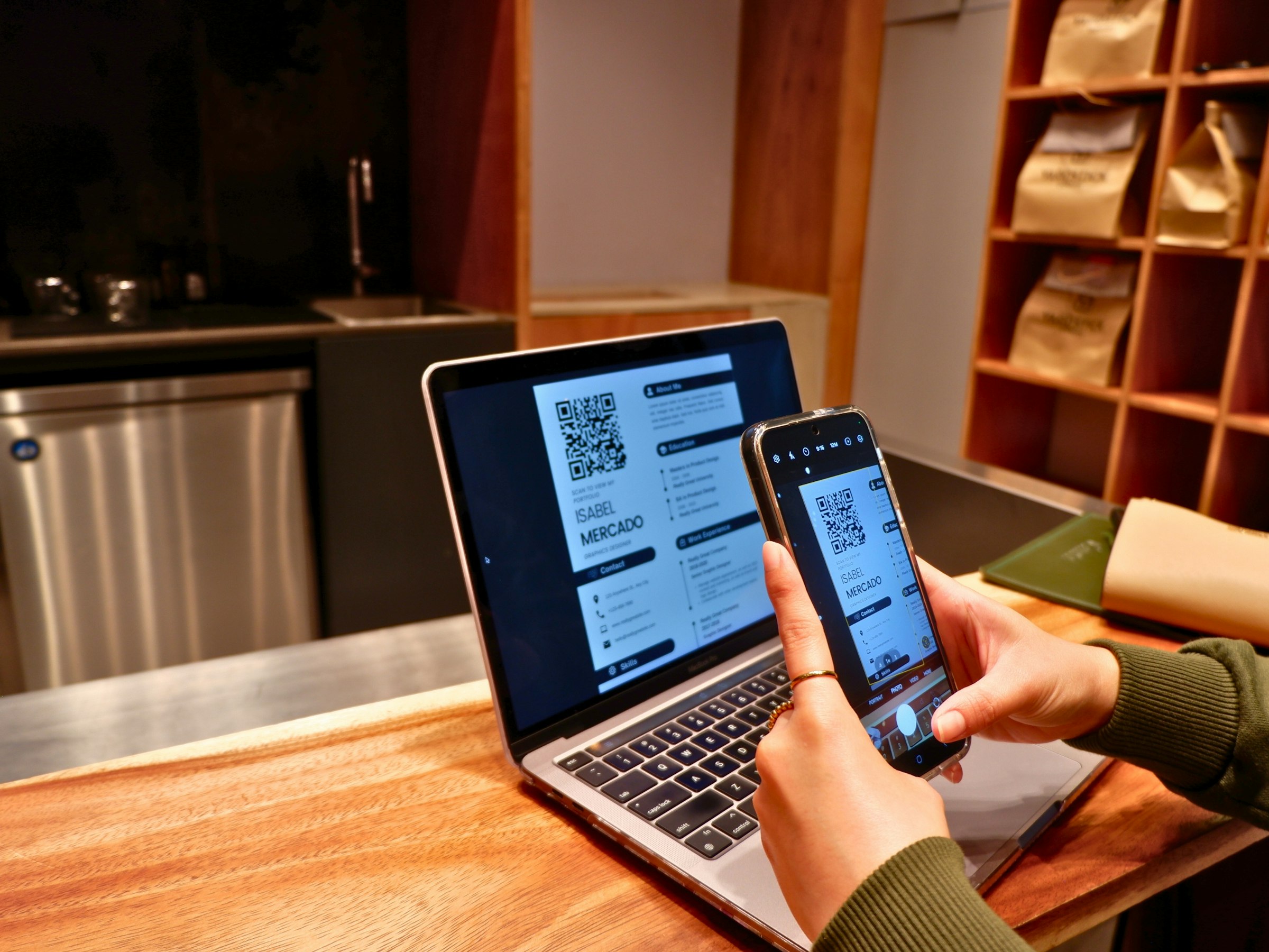 Person scanning a QR code on a phone with a laptop open behind it (moving between devices in the same session)
